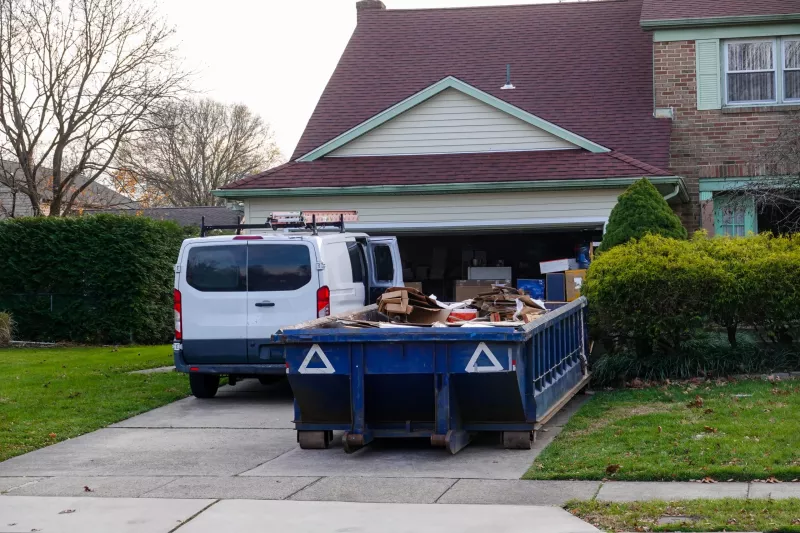 Low blue dumpster full of cardboard and construction debris in front of a full garage and next to a white worker's van